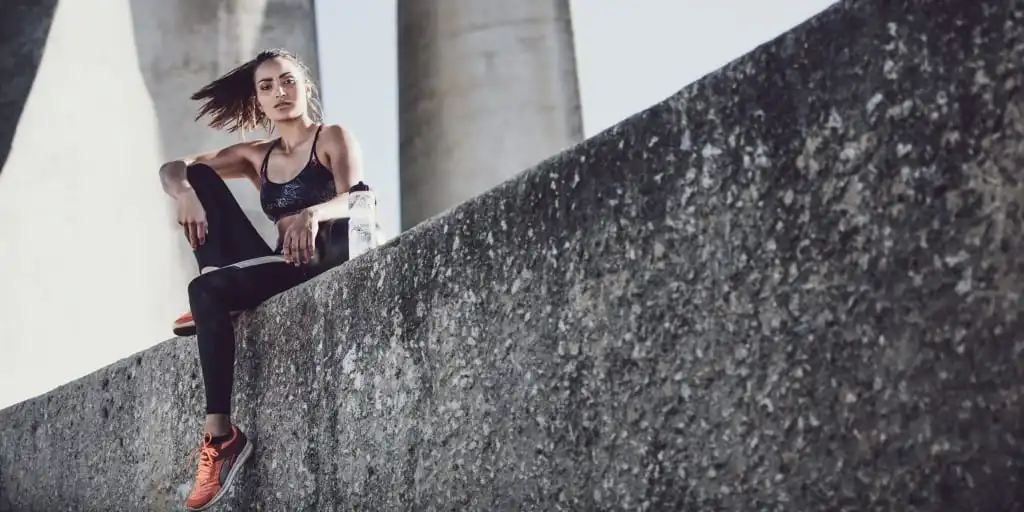 Drinking Water Benefits by Hydro Coach, a woman, her long brown hair in the wind, after workout sitting on top of a wall having a water bottle in front of her.